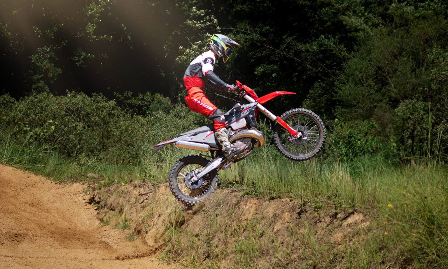 Piloto na MXF 250 TSX em um salto alto, após subir rampa de terra em trilha com fundo de vegetação.