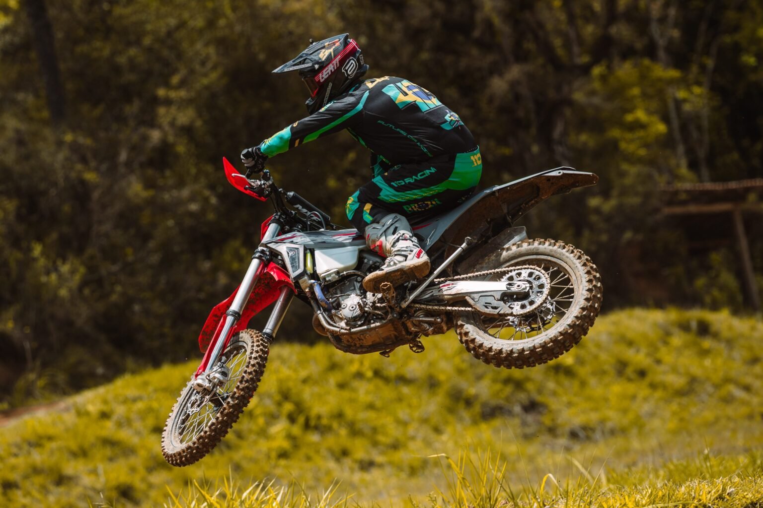Piloto (roupa verde e preta) em salto lateral com a MXF 250 RXi sobre grama e terra.