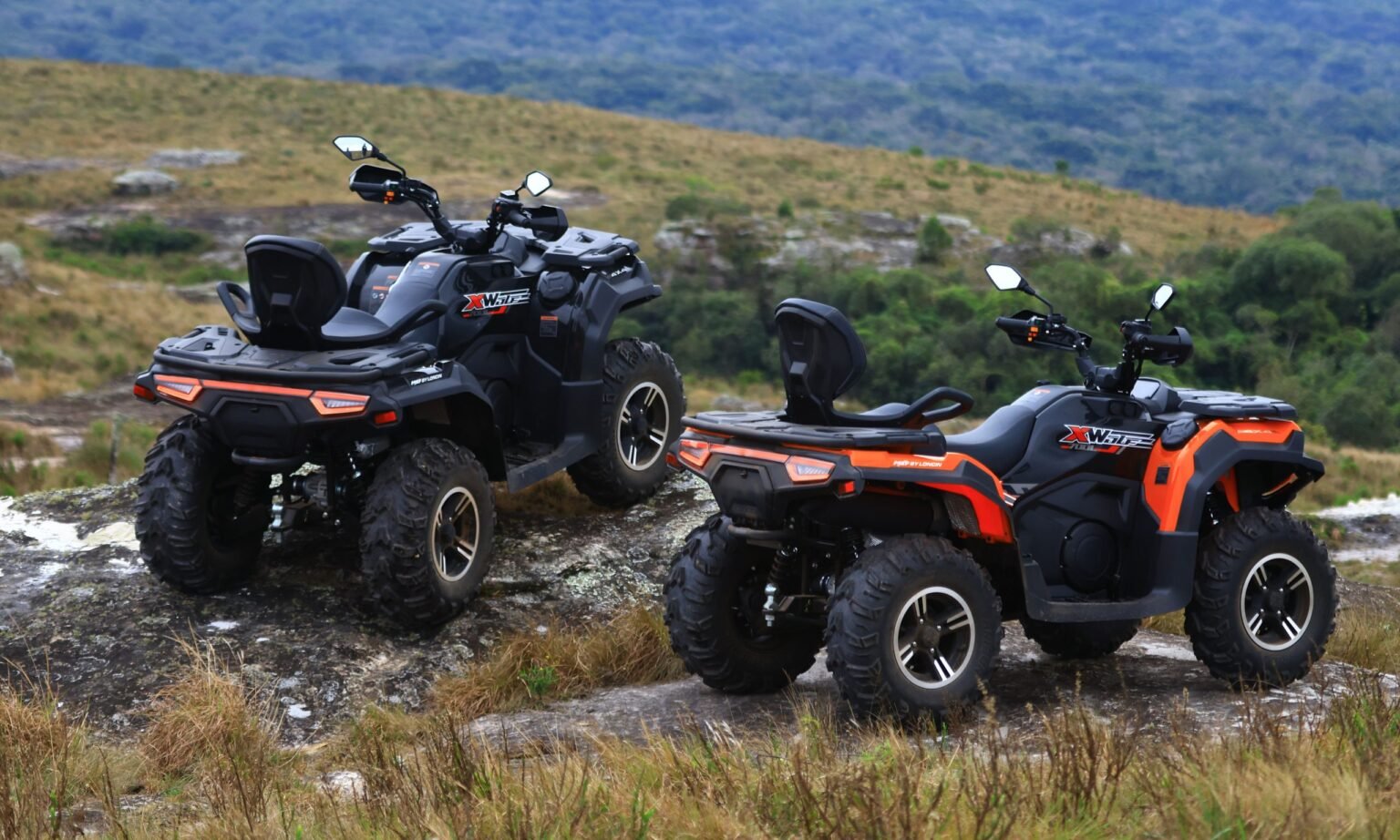 Dois quadriciclos MXF Wolf 700L, um na cor preta e outro laranja, em um cenário de montanhas, destacando o design robusto e os acessórios de série na MXF Burt Motos em Campinas.